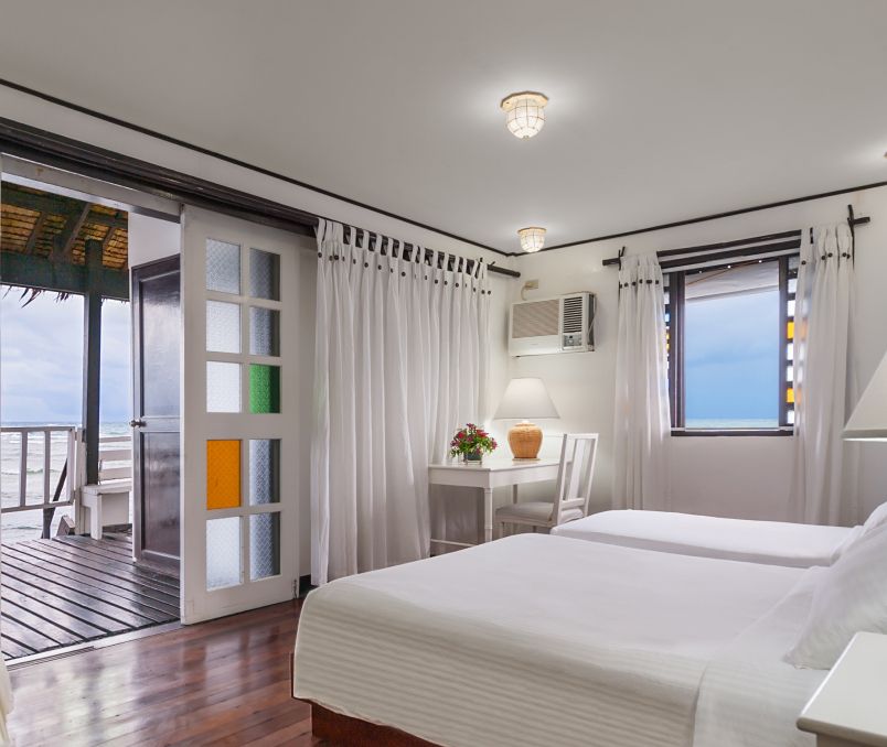 Bedroom with two white beds, a desk, and a balcony overlooking the ocean.