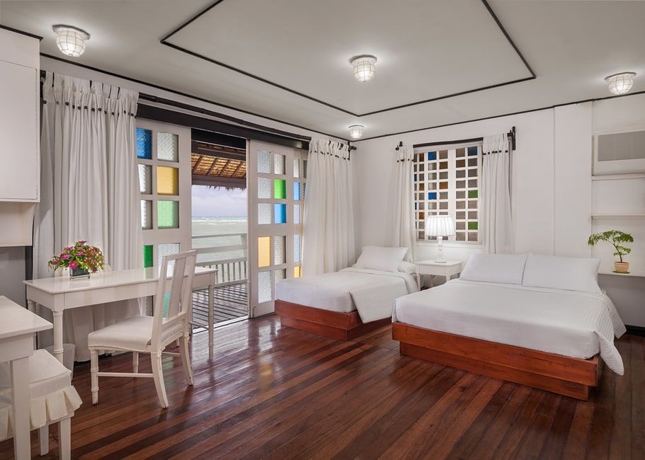 Bedroom with two beds, desk, and balcony access with colorful window panes, overlooking a water view.