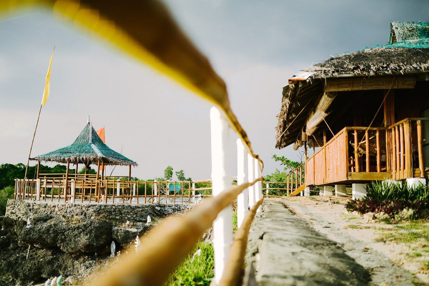 A bamboo railing leads to a gazebo and a wooden hut on a rocky cliff, under a cloudy sky.