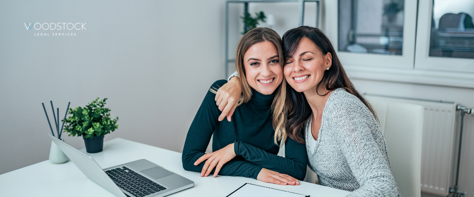 Two smiling women embracing at a modern office desk, with a laptop and documents in front of them