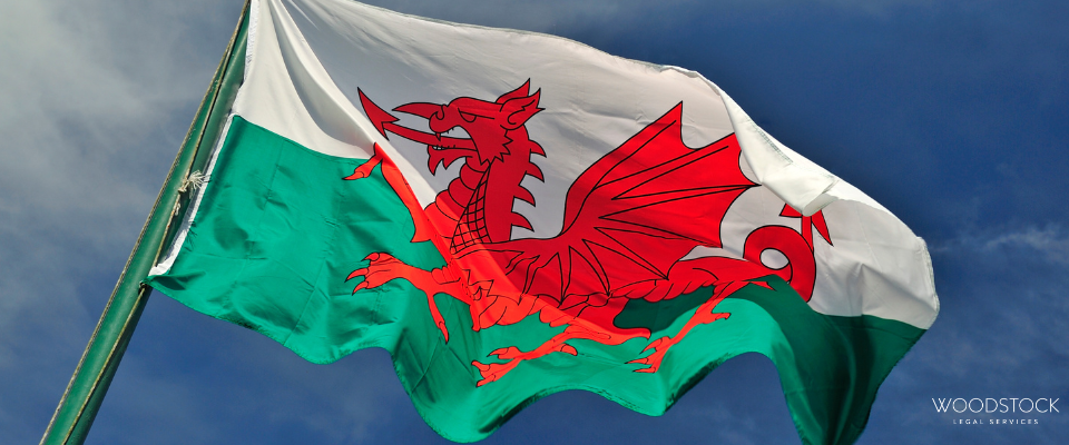The Welsh flag waving against a dark blue sky, showing a red dragon on white and green with a Woodstock Legal Services logo in the bottom right corner.