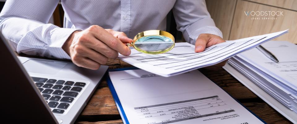 Person reviewing legal and financial documents with a magnifying glass at a desk, laptop and paper