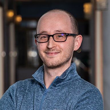 A bald man wearing glasses and a blue sweater is smiling for the camera.