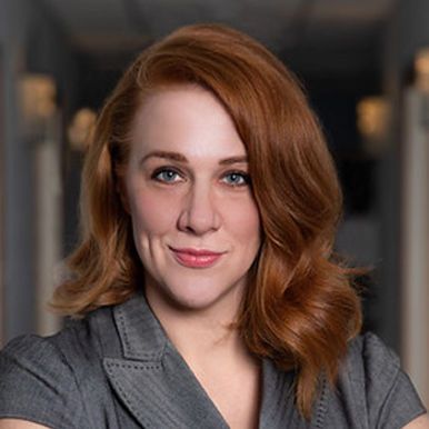 A woman with red hair and blue eyes is wearing a gray jacket and smiling.