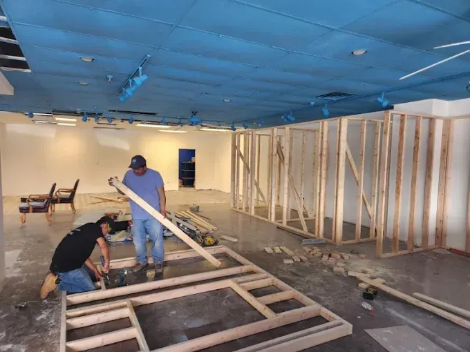Two workers construct a wooden wall frame inside a building with a blue ceiling and white walls.