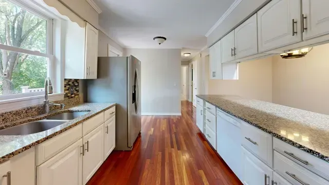 A white kitchen with speckled granite countertops, stainless steel refrigerator, and hardwood floors.