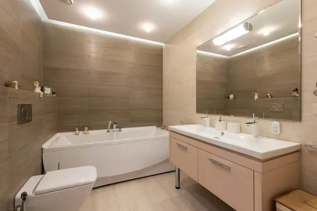 A modern, neutral-toned bathroom featuring a white bathtub, a wall-mounted toilet, and a vanity with a large mirror.