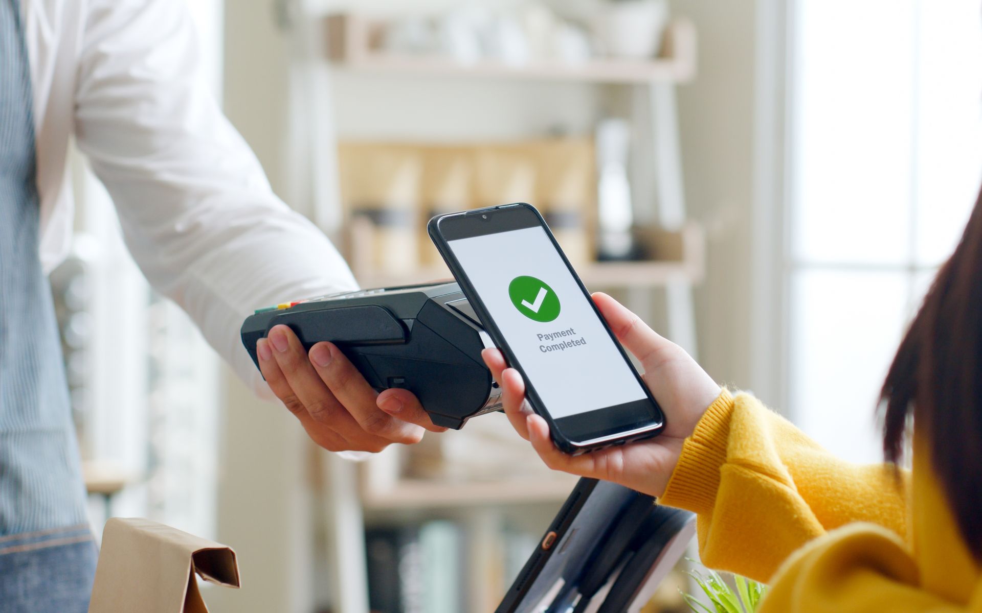 Person taps phone to card reader while another holds a smartphone with a green checkmark on screen.