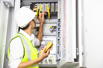 An electrician is working on an electrical box while holding a multimeter.