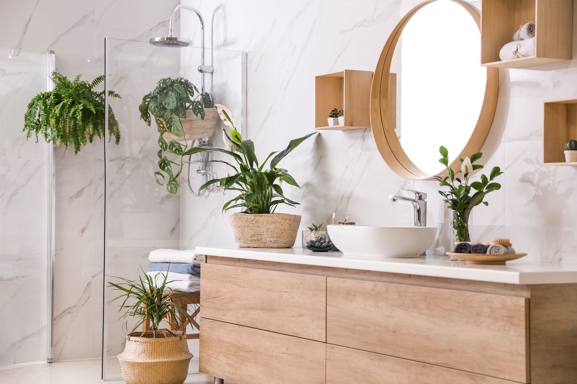 Bathroom with a wooden vanity, round mirror, and several potted plants. White marble walls and glass shower.