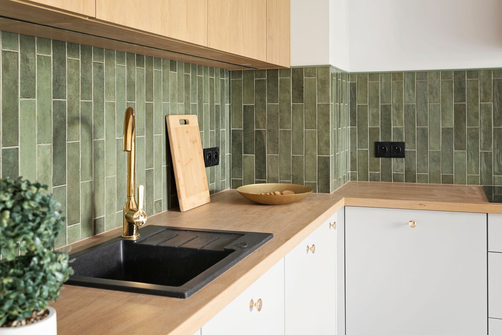 Kitchen corner with olive green tiled backsplash, gold faucet, wooden countertops, and white cabinets.