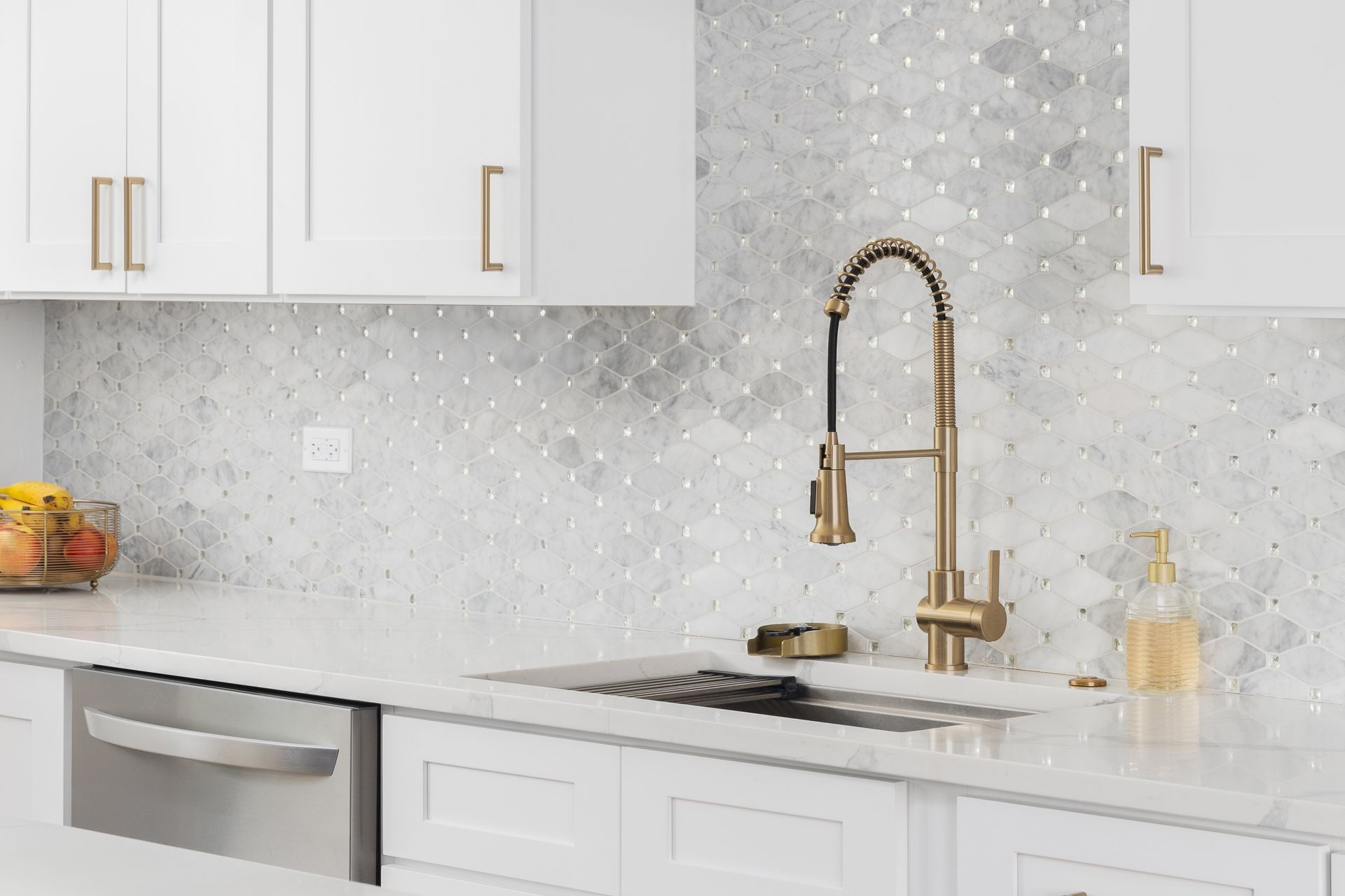 A modern kitchen sink with a gold faucet, white cabinets, white countertops, and a gray-and-white patterned tile backsplash.