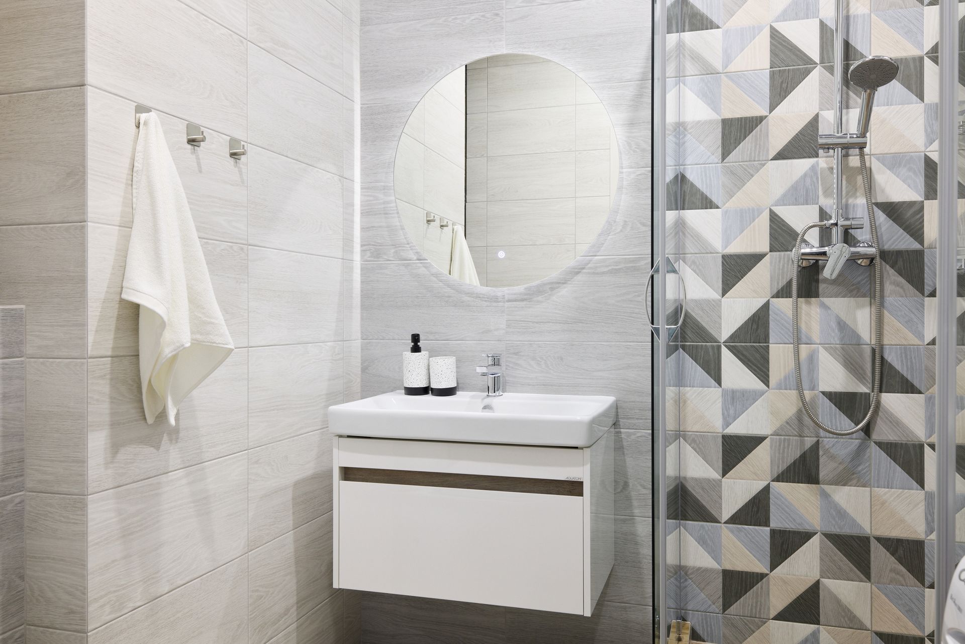 Modern bathroom with white vanity, geometric tile shower, and round mirror.
