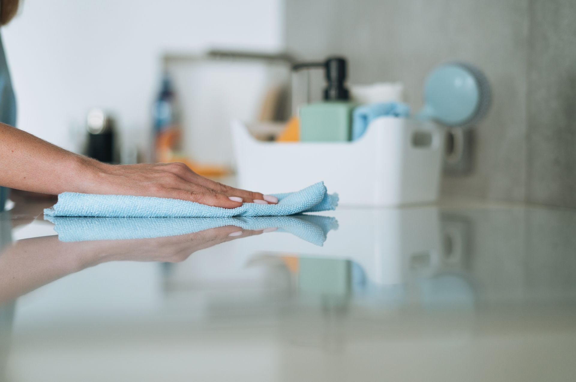 Person wiping a light-colored countertop with a blue cloth, items in white bin in background.