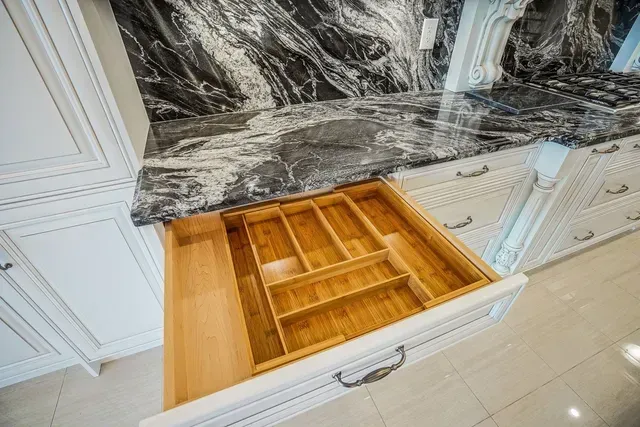 Open kitchen drawer with bamboo utensil organizer, below dark granite countertop. White cabinets.