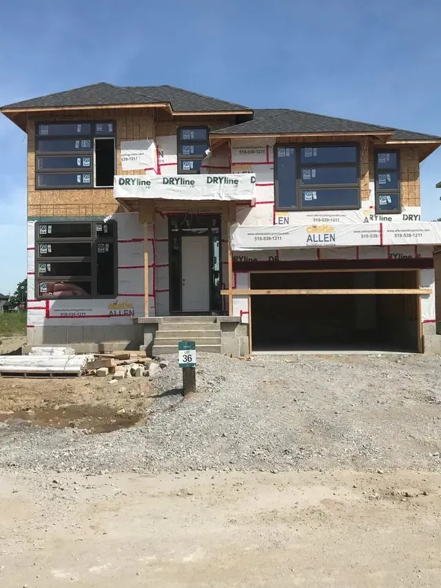 A two-story house under construction with large windows and a gravel driveway.