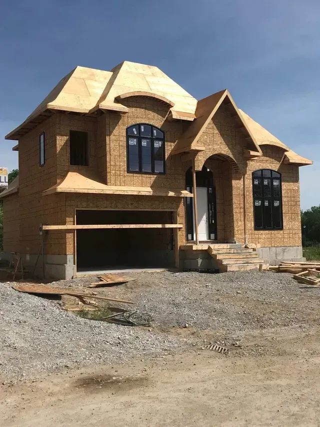 House under construction; wood frame with visible plywood siding, garage, windows, and front steps.