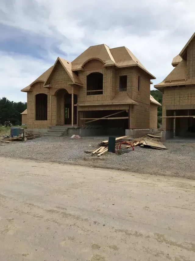 House under construction, unfinished wood exterior, two-story with attached garage, gravel driveway.