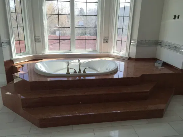 Built-in bathtub on a raised platform with steps, in front of a large window, brown tones.