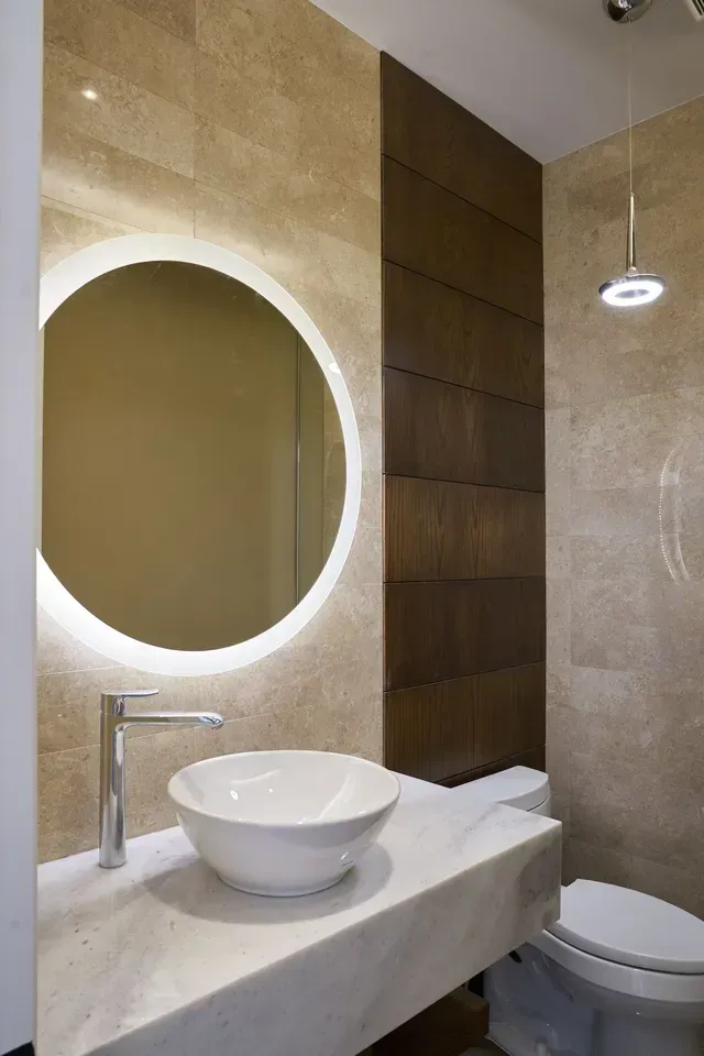 Modern bathroom with round mirror, vessel sink, and toilet, featuring neutral tones and recessed lighting.