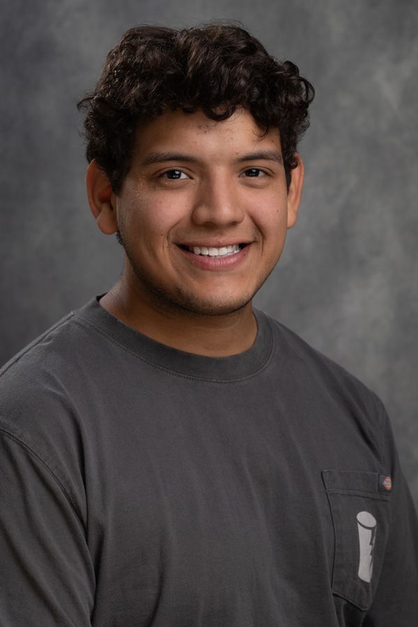 A young man with curly hair is wearing a gray shirt and smiling for the camera.