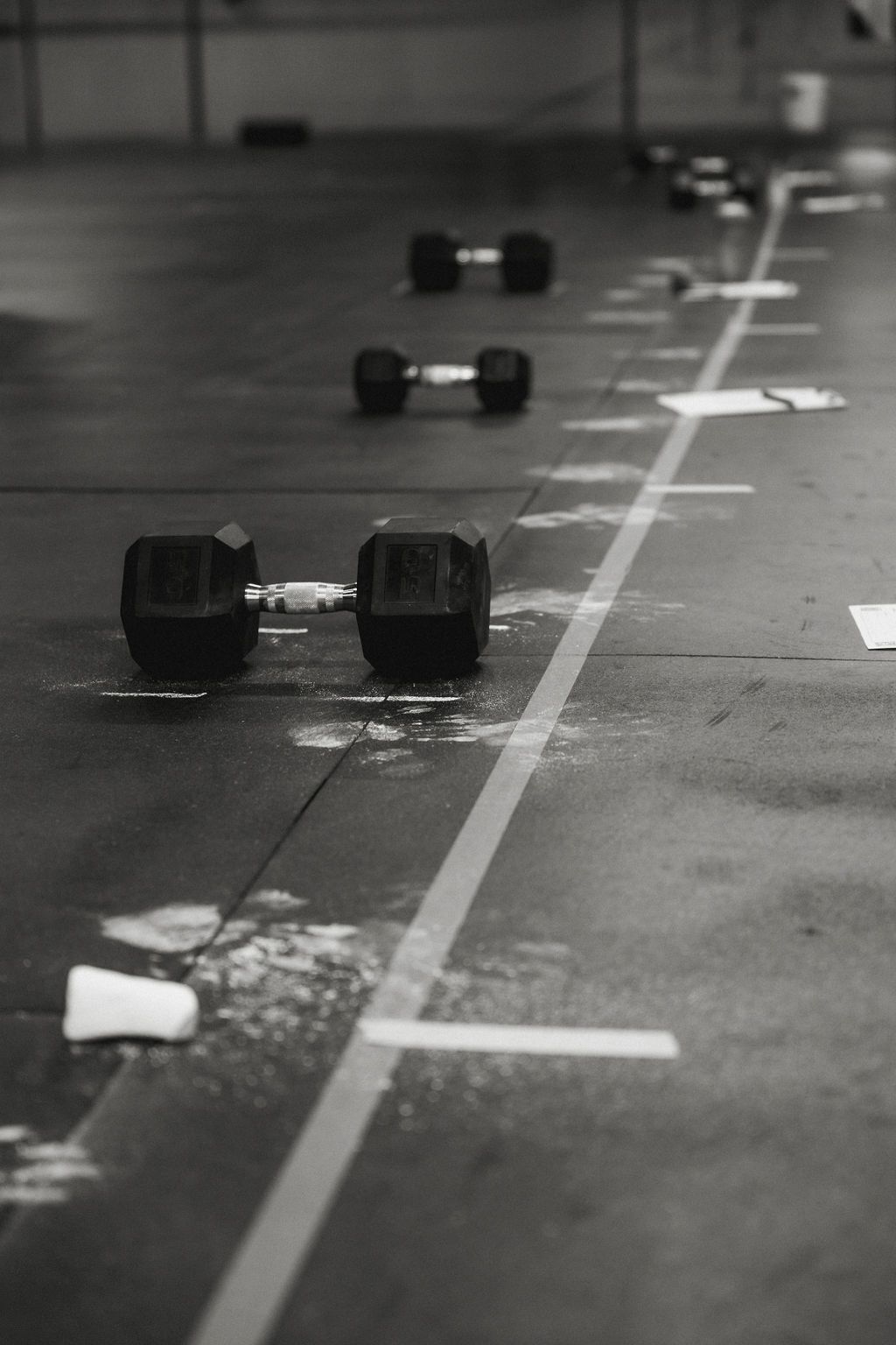 Row of dumbbells on a gym floor with white lines and markings, in black and white.