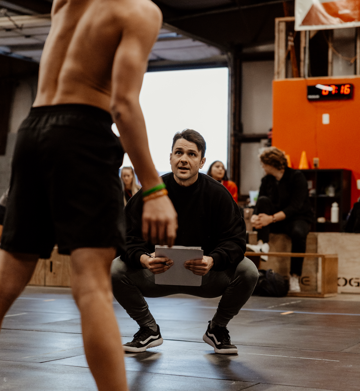 Man in black sweats watches athlete, squatting, in a gym in Spanish Fork, UT.