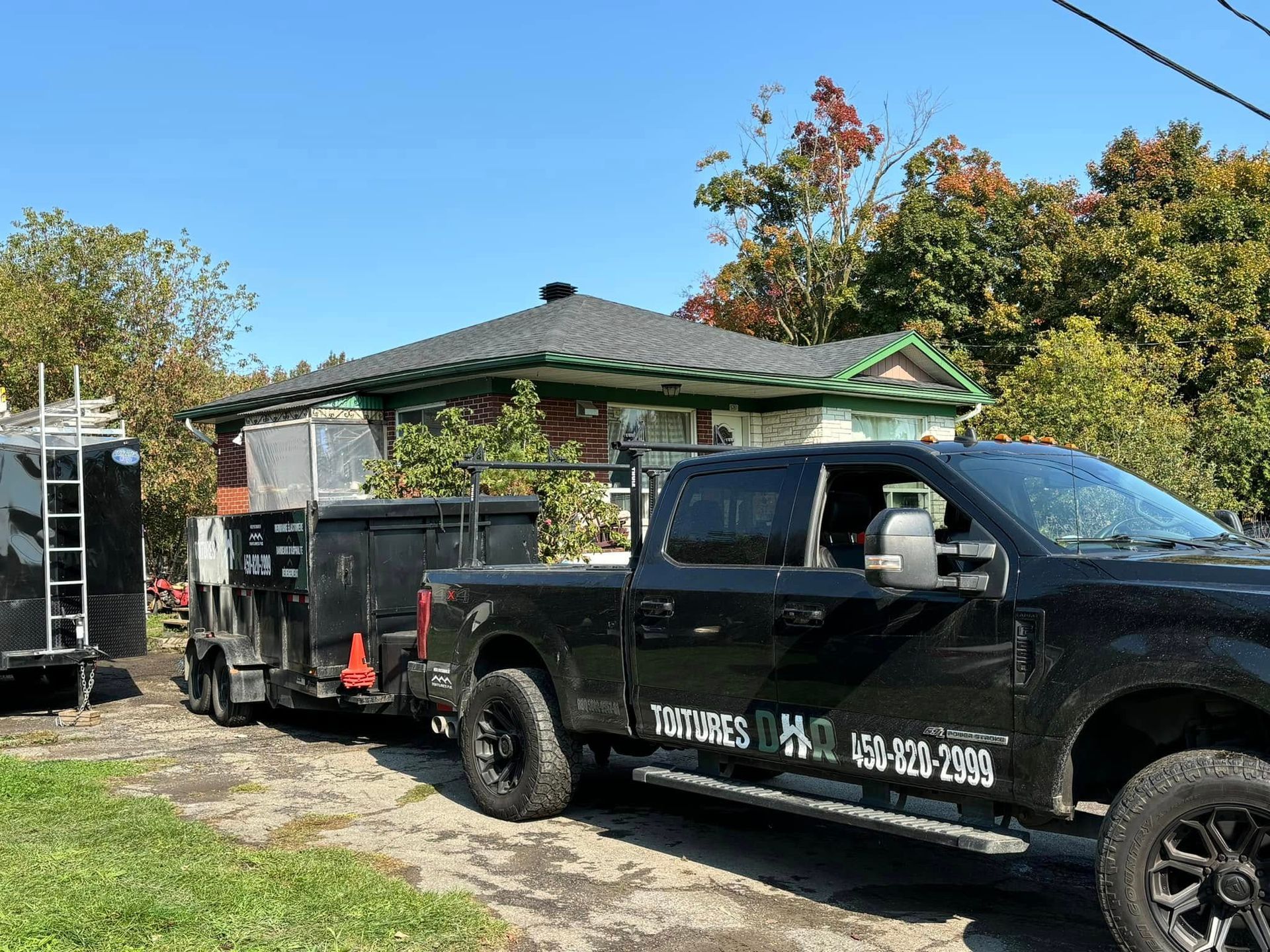 Un camion noir avec remorque est stationné devant une maison, probablement pour des travaux d'aménagement paysager ou de construction, sous un ciel bleu.