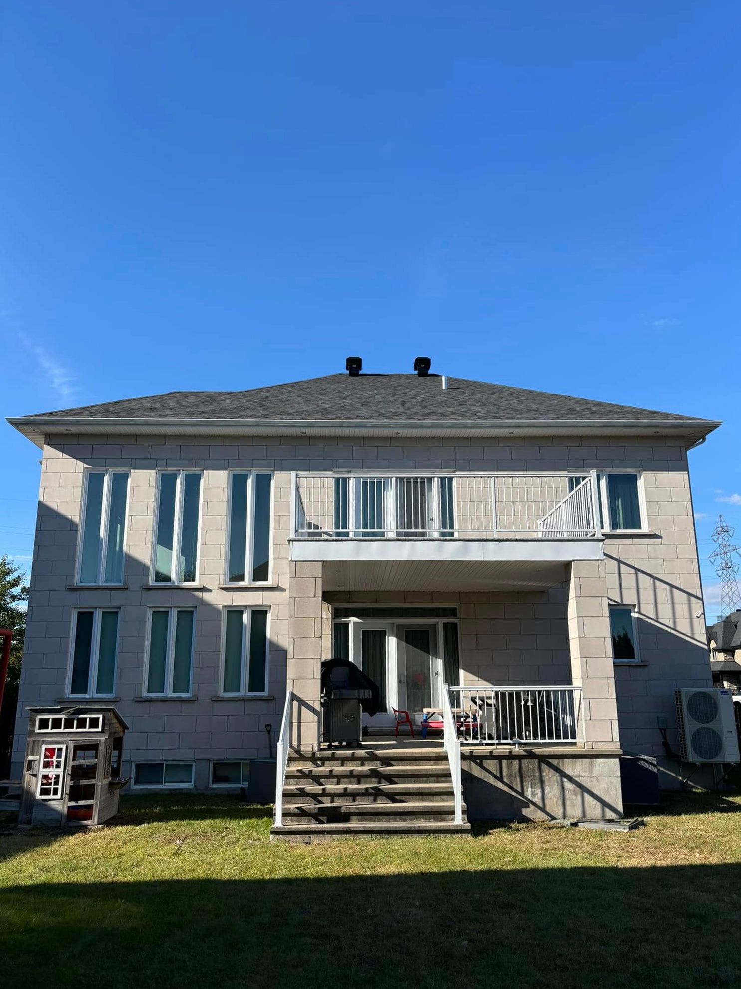 Maison grise de deux étages avec de nombreuses fenêtres et un balcon, située sur une pelouse verdoyante sous un ciel bleu azur.
