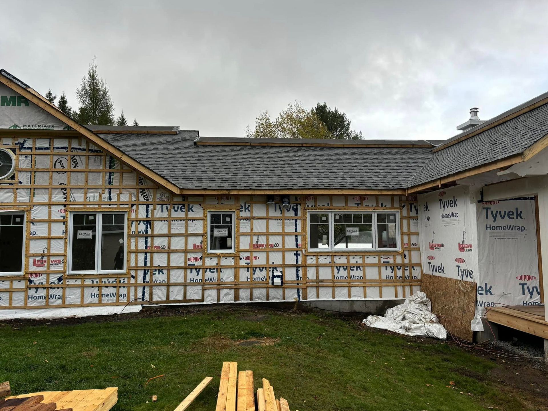 Vue extérieure d'une maison en construction ; charpente en bois, fenêtres et bâche Tyvek visibles. Toit gris.
