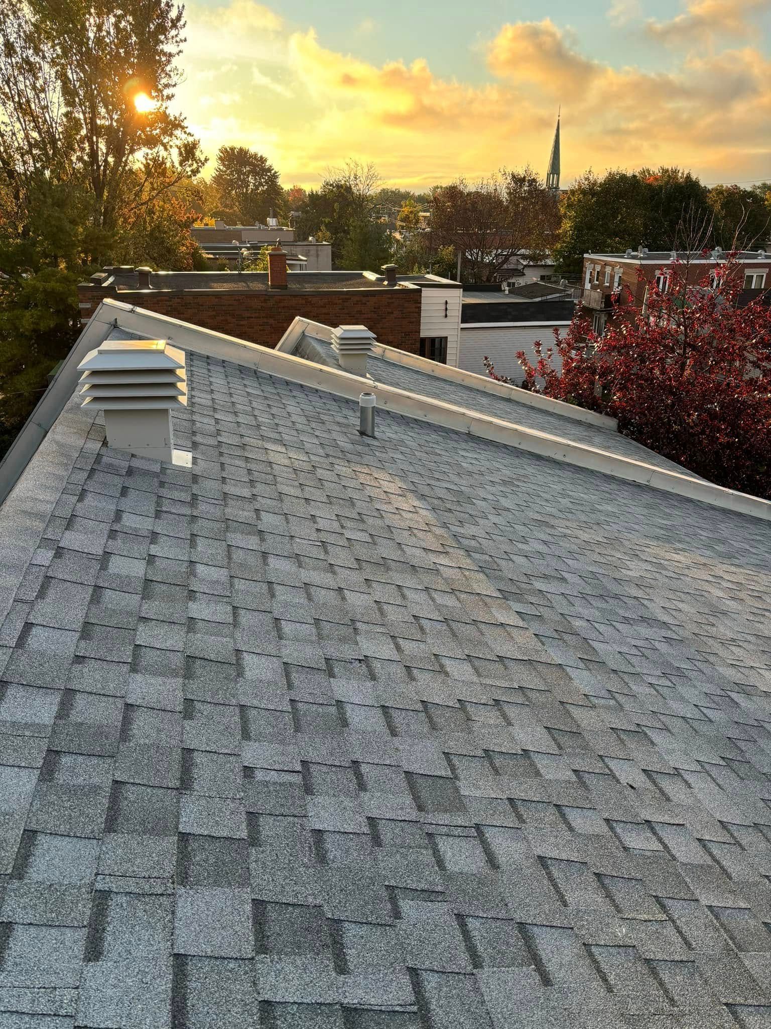 Toiture en bardeaux gris se détachant sur un ciel de coucher de soleil ; les conduits d'aération et les bâtiments environnants sont visibles.