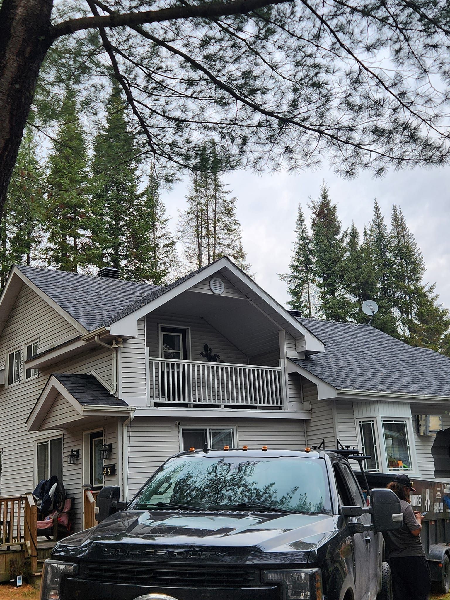 Maison à deux étages avec un camion noir garé devant. Bardage blanc, toit foncé, arbres en arrière-plan. Ciel couvert.