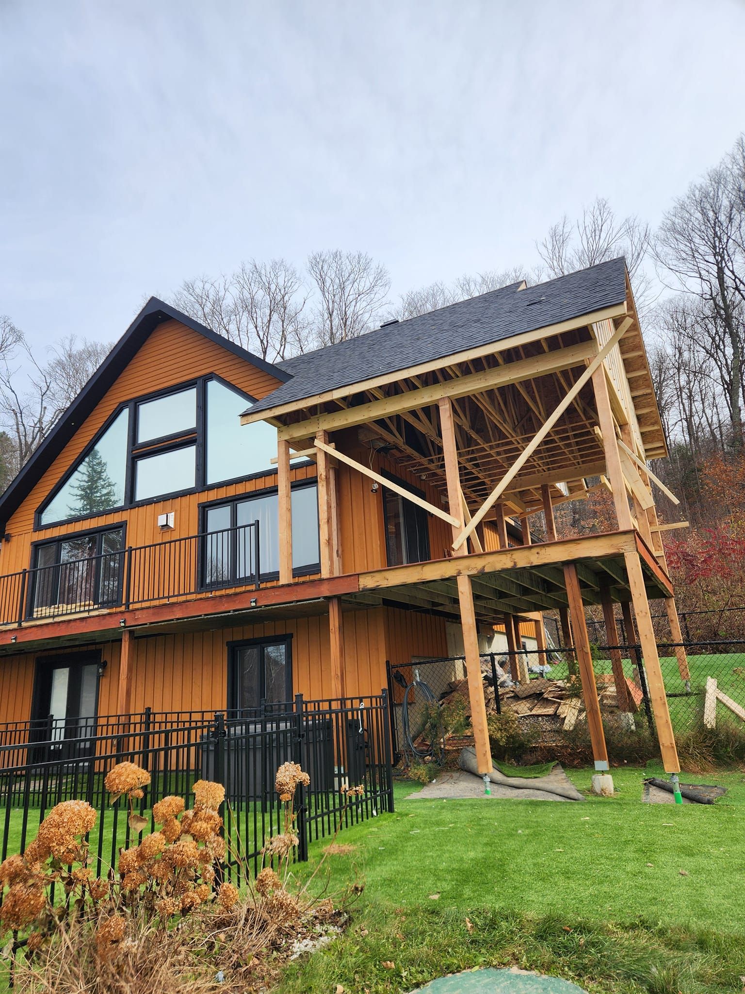Maison en bois de deux étages avec une terrasse en construction, adossée à une colline et sous un ciel nuageux.