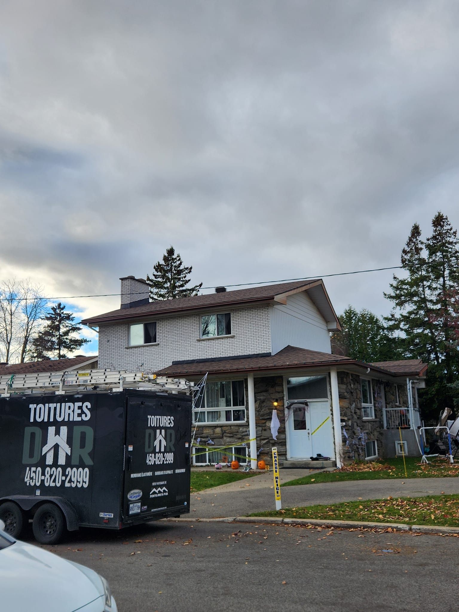 Maison à deux étages avec façade en pierre, toiture en cours de réparation. Une remorque portant l'inscription « Toitures DHR » est garée devant.
