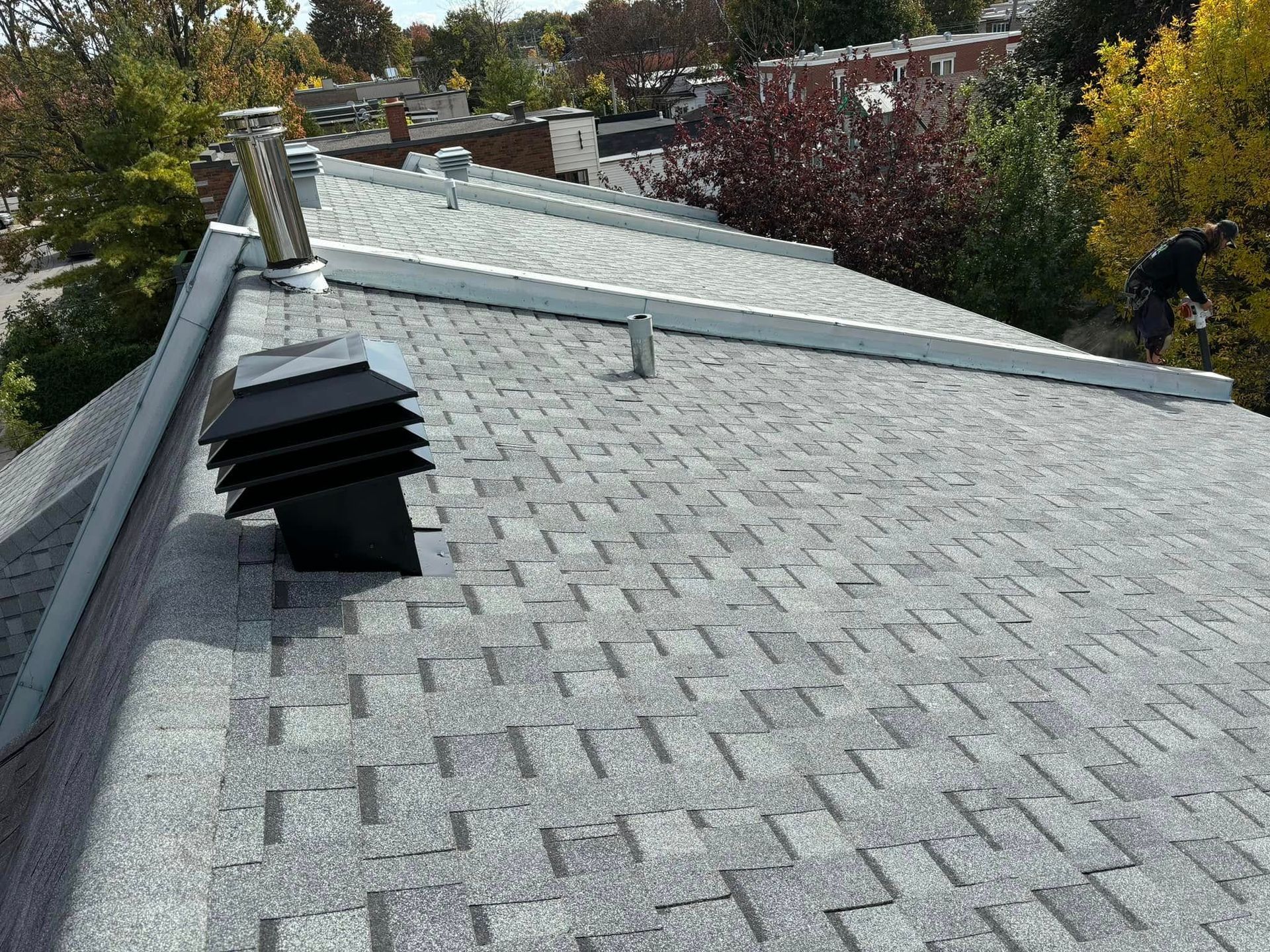 Toiture en bardeaux d'asphalte gris avec aérations, solins métalliques et arbres en arrière-plan.