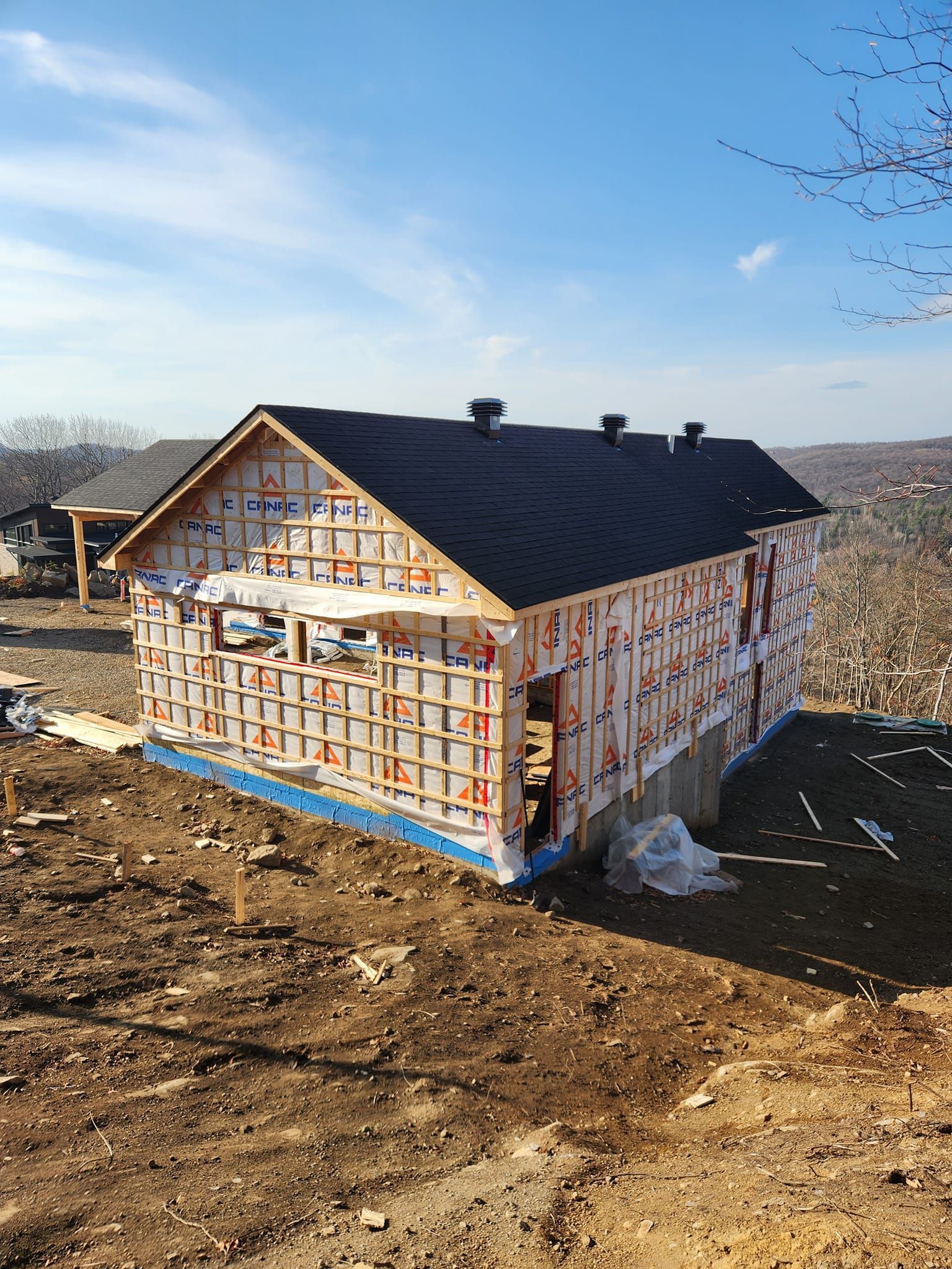 Construction d'une maison au toit de bardeaux noirs, entourée de terre par une journée ensoleillée.