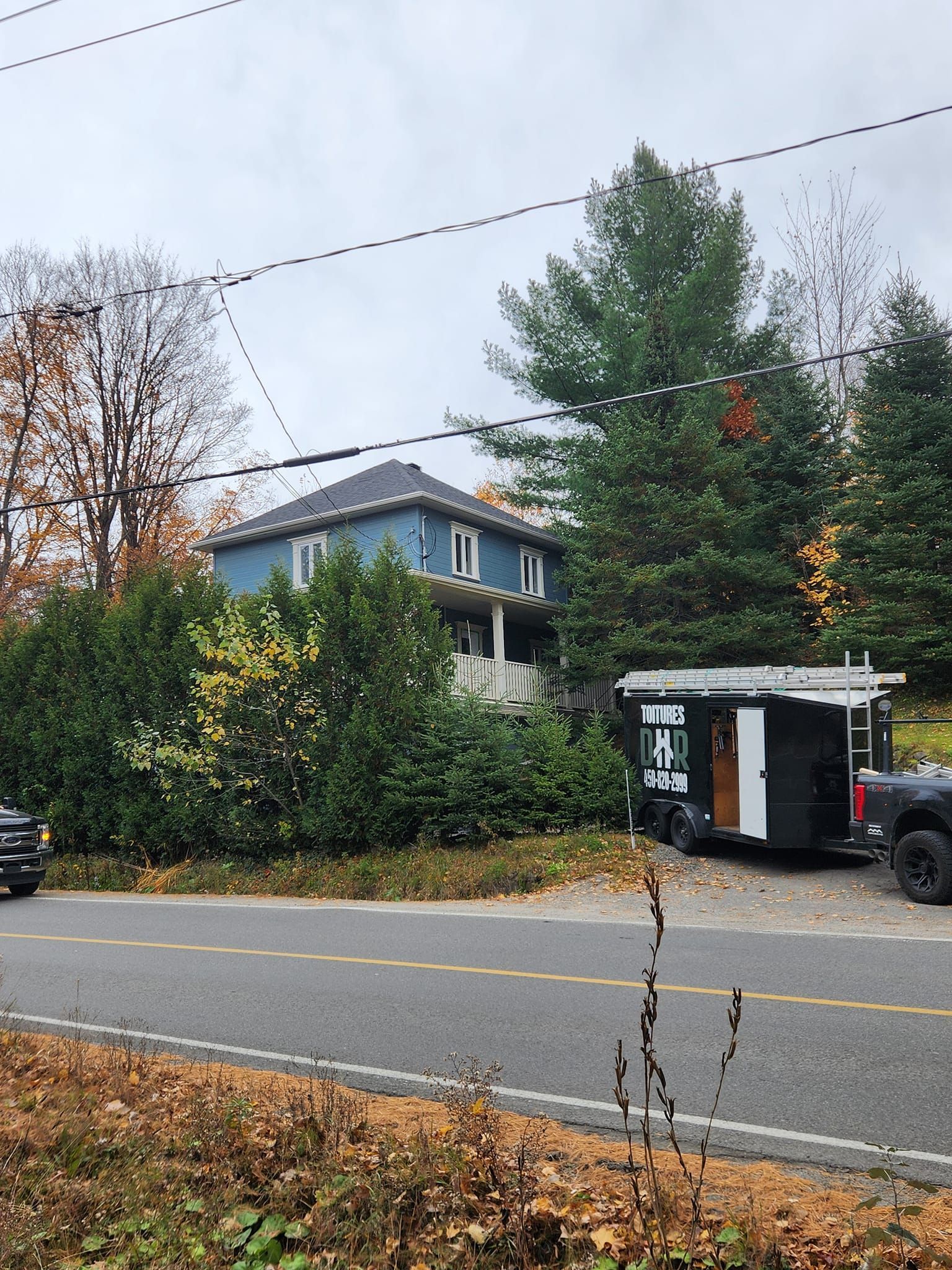 Maison bleue derrière des buissons, caravane garée en bord de route, feuillage d'automne, ciel couvert.