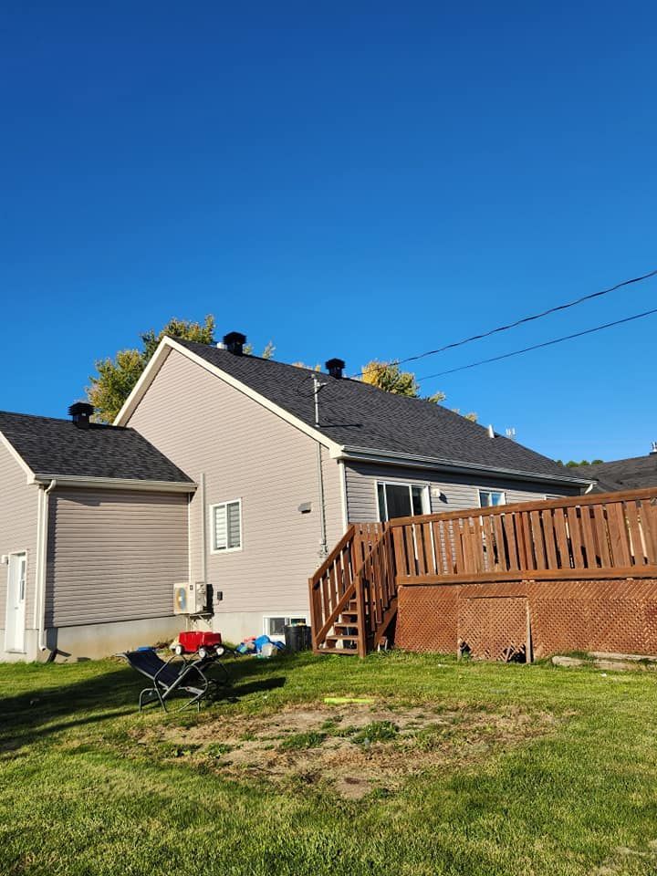 Maison beige avec toit gris foncé et terrasse marron sous un ciel bleu azur.
