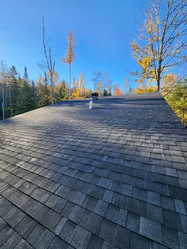 Toit en bardeaux gris avec cheminée et conduits de ventilation, sous un ciel bleu et des arbres d'automne.
