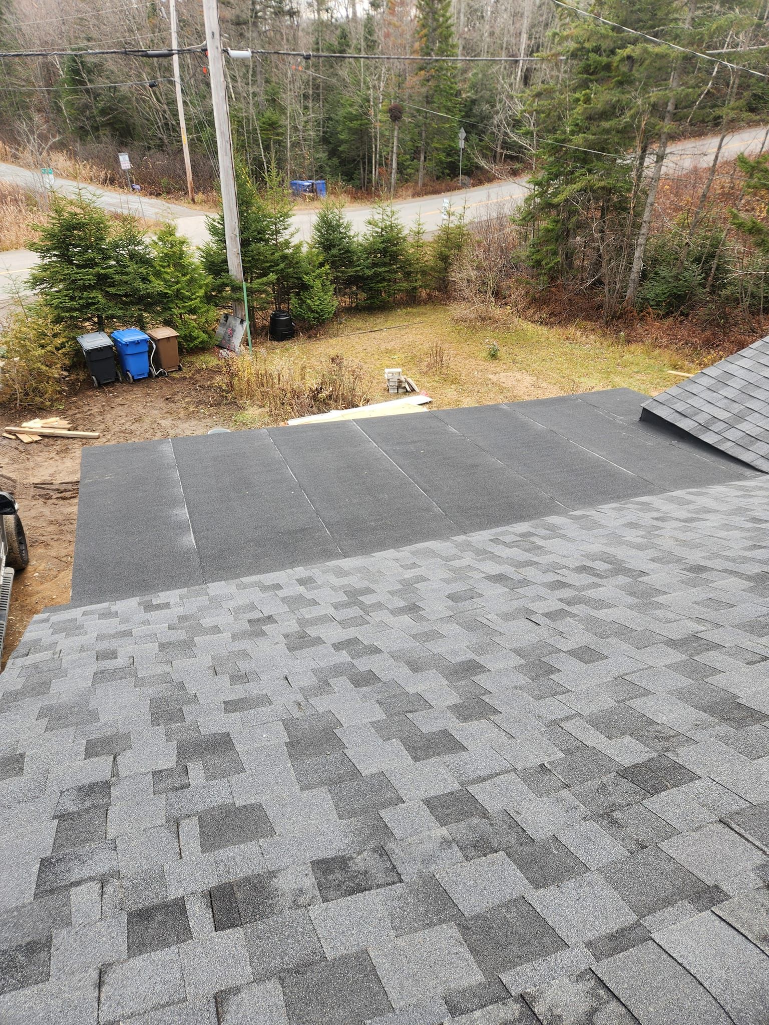 Pose de bardeaux gris foncé sur le toit. Vue sur le jardin, les arbres et le poteau électrique.