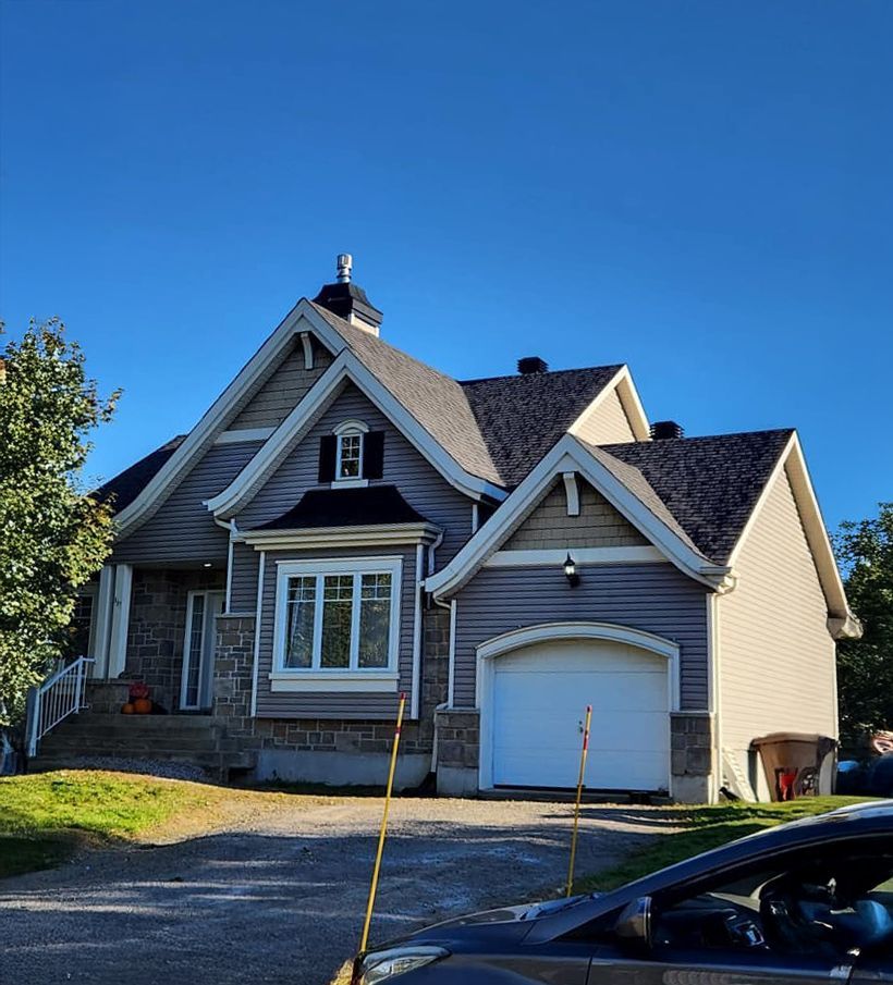 Maison grise et beige avec façade en pierre et porte de garage blanche, sous un ciel bleu azur.