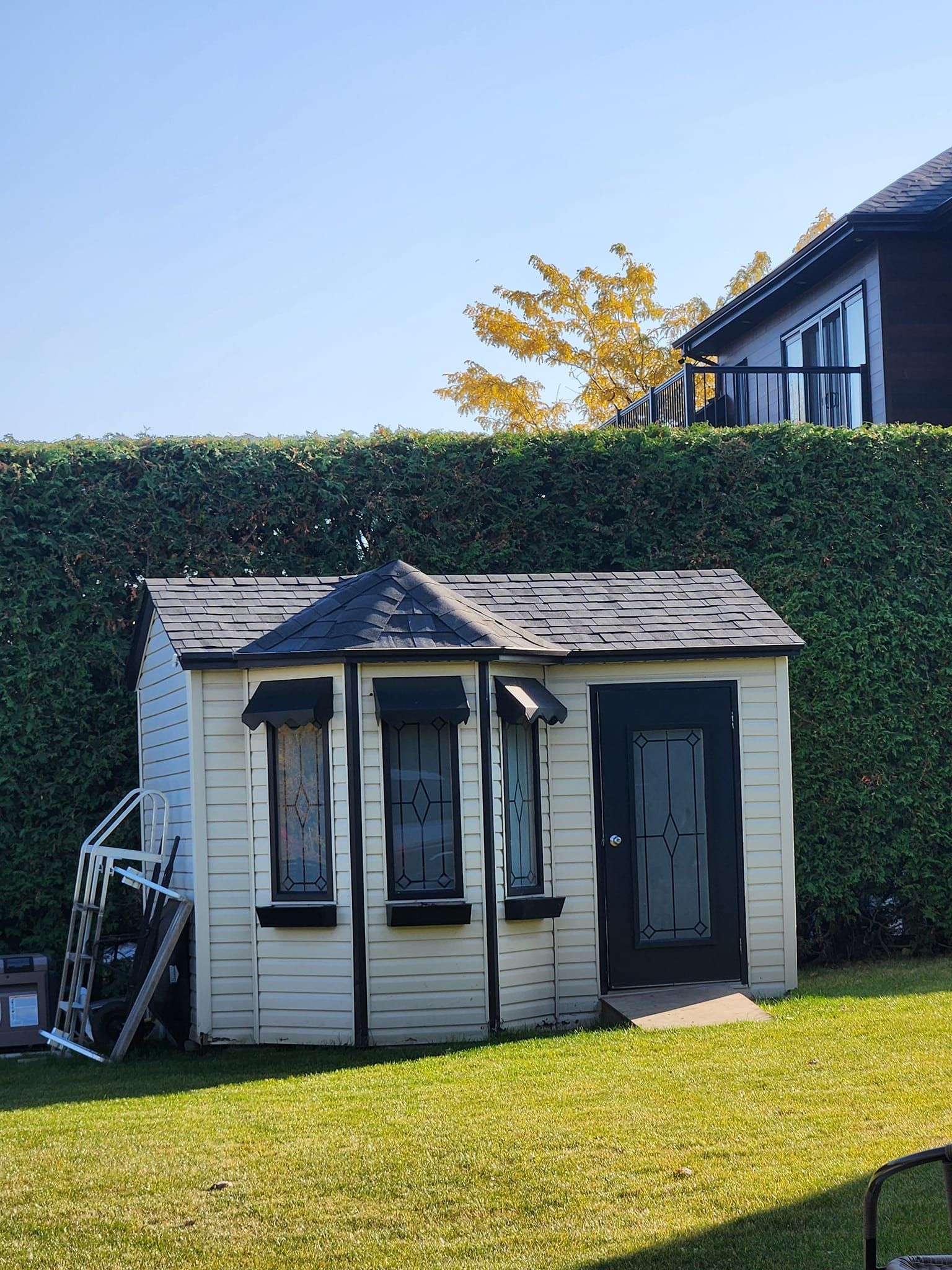 Maisonnette blanche aux finitions noires, dans un jardin gazonné, avec une haute haie et une maison en arrière-plan.