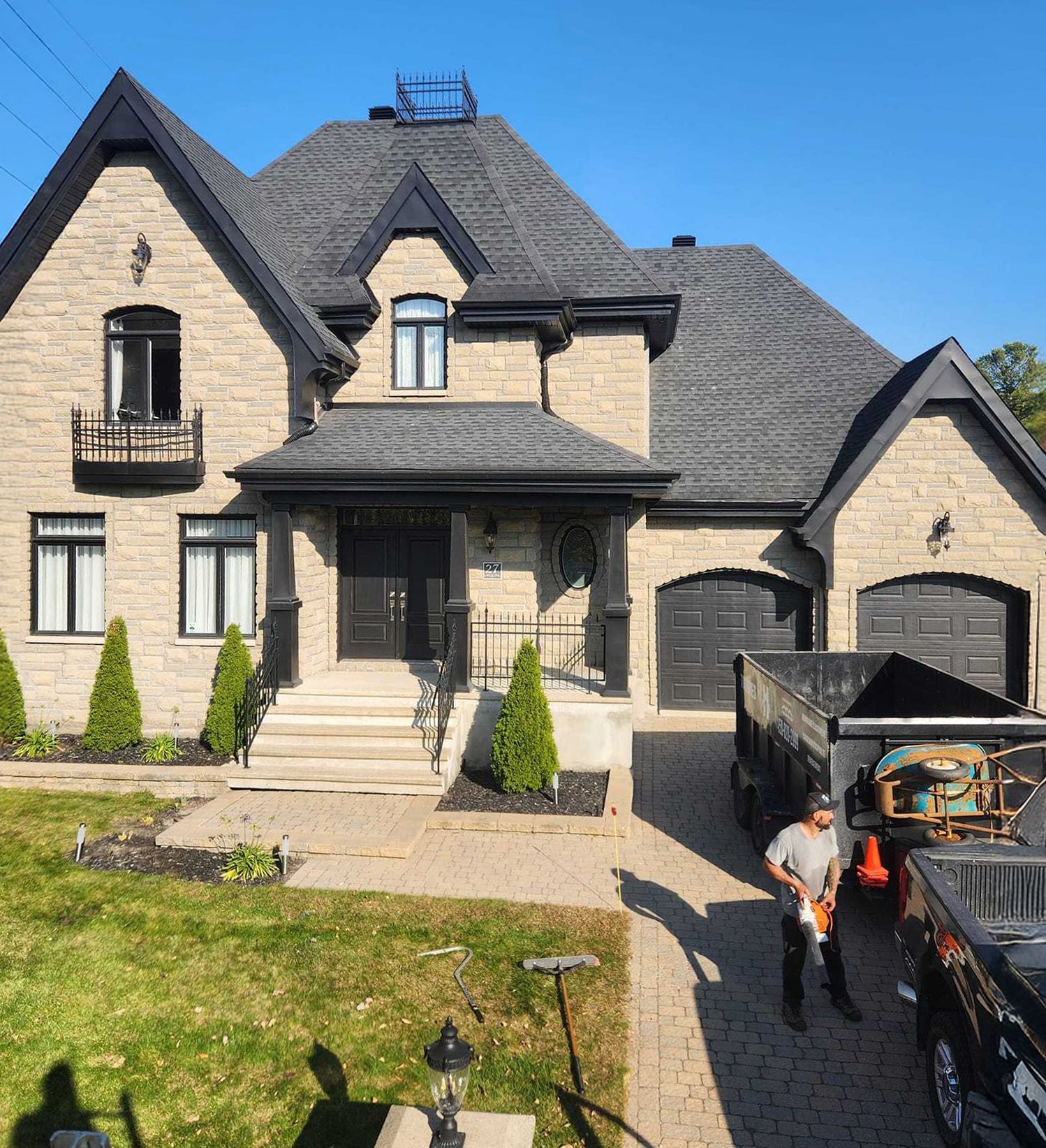Maison à façade en pierre, toit sombre et portes de garage ; ouvrier avec des outils près d'un camion sur une allée pavée.