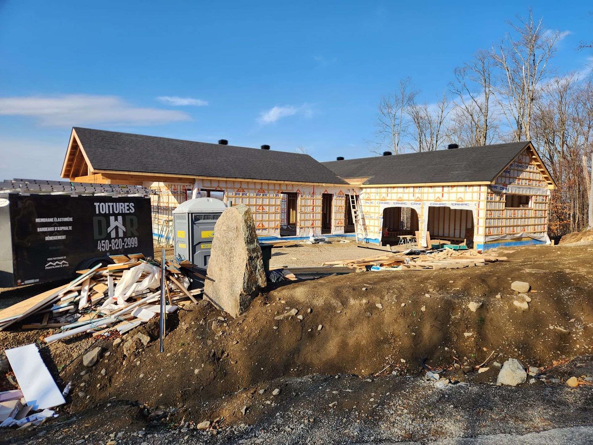 Chantier de construction avec une maison en construction, une benne à ordures et des toilettes portables sous un ciel bleu.