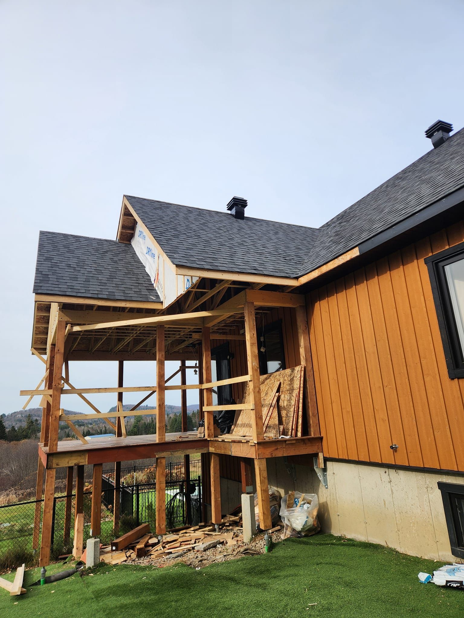 Maison avec terrasse en bois partiellement construite et charpente apparente ; bardage brun, toit noir.