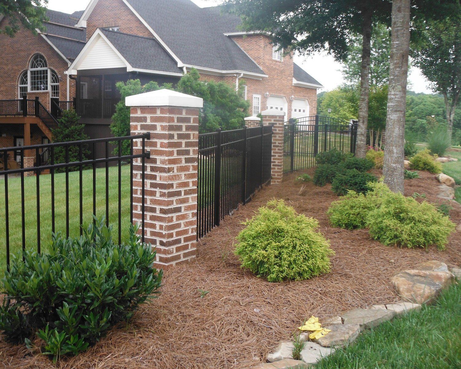 A black fence surrounds a brick pillar in front of a large brick house