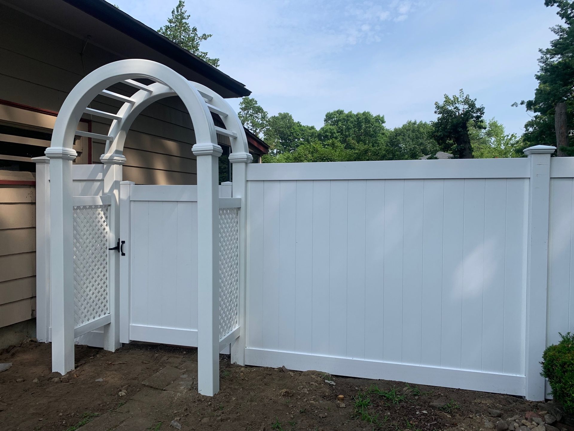 White vinyl fence with arched gate, leading to yard with trees.