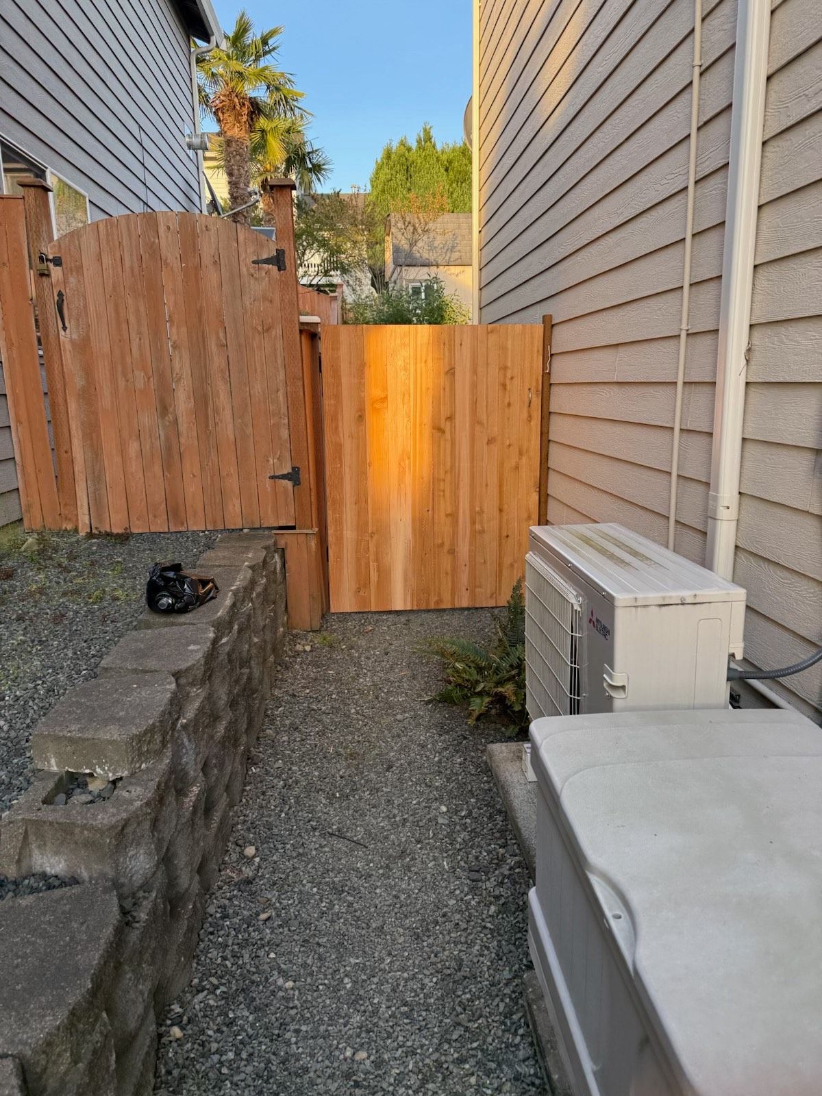 Wooden gates in a narrow passage between a house and a retaining wall. Gravel path. Sunlight.