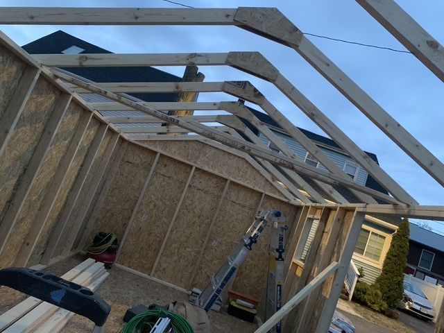 Wooden shed under construction, with roof trusses and wall framing visible outdoors.