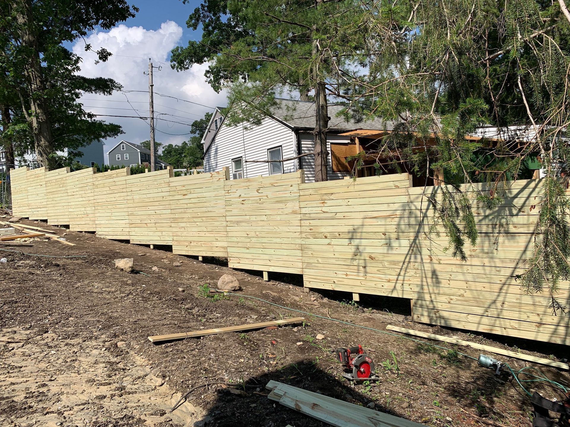 A long, light-colored wooden fence being built outdoors near a house and trees.